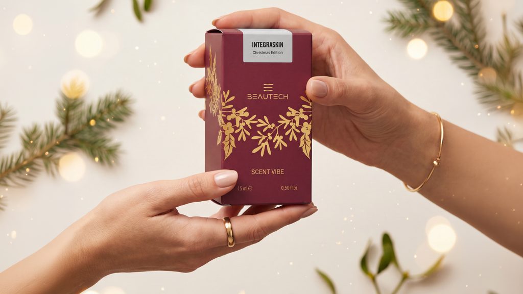 Due mani tengono la confezione Scent Vibe Integraskin Christmas Edition di Beautech, color bordeaux con decorazioni dorate, tra rami di pino e luci natalizie.